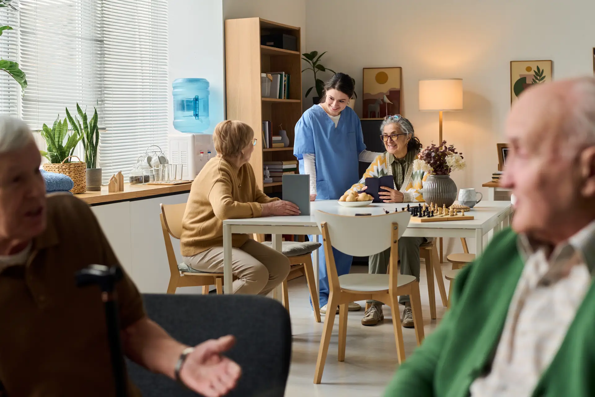 A caregiver with two residents in a common area, a chess set and books on the table